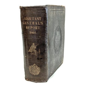 Puede incluir: Un libro antiguo de color marrón oscuro, titulado "ADJUTANT GENERAL'S REPORT 1864." La portada presenta letras doradas e ilustraciones en relieve. El lomo del libro muestra desgaste. El libro está sobre un fondo blanco.