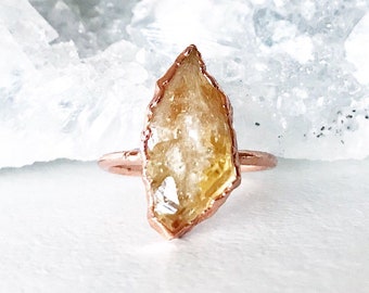 May include: A rose gold ring with a raw, yellow citrine stone set in the center. The stone is irregularly shaped and has a natural, rough texture.