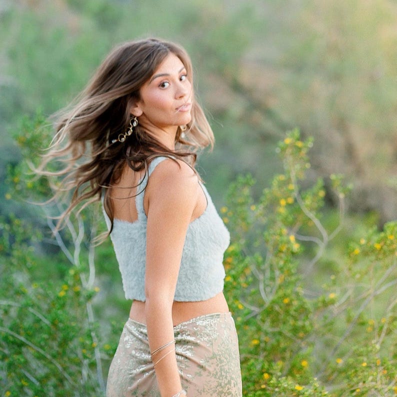 May include: A woman wearing a light blue crop top and a gold skirt poses outdoors. Her hair is blowing in the wind, and she is wearing earrings and bracelets. The background is a blurred view of green foliage and yellow flowers.