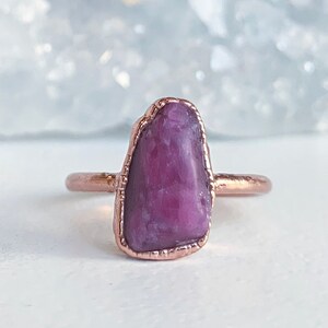 Puede incluir: Un anillo de color cobre con una gema grande, de forma irregular, de color rosa intenso. La gema est&aacute; engastada en un bisel de cobre y tiene un aspecto pulido y natural. La banda del anillo es una banda de cobre fina y sencilla.