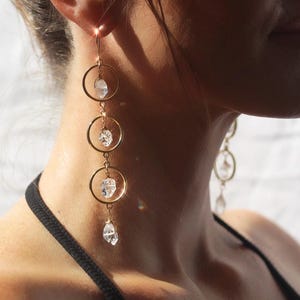 May include: Gold-tone earrings with three connected hoops, each holding a clear, irregularly shaped crystal. The earrings hang from a hook and are shown against a blurred background. The jewelry is a delicate accessory.