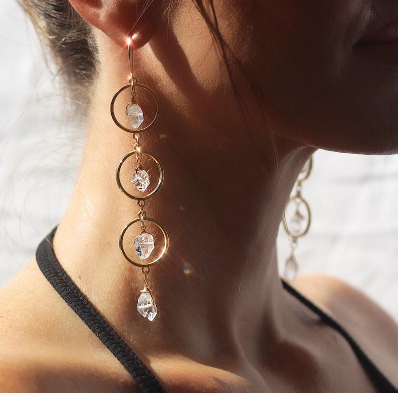 May include: Gold-tone earrings with three connected hoops, each holding a clear, irregularly shaped crystal. The earrings hang from a hook and are shown against a blurred background. The jewelry is a delicate accessory.