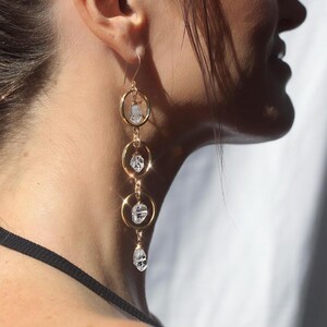 May include: Gold-tone earrings with a cascading design. Each earring features three gold hoops connected by delicate chains. Clear, faceted crystals are suspended within each hoop, creating a sparkling effect. The earrings are designed to dangle and catch the light.