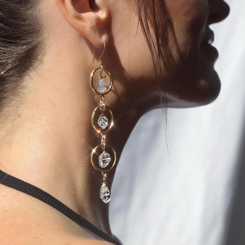 May include: Gold-tone earrings with a cascading design. Each earring features three gold hoops connected by delicate chains. Clear, faceted crystals are suspended within each hoop, creating a sparkling effect. The earrings are designed to dangle and catch the light.