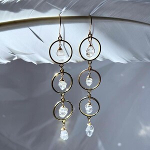 May include: Gold-tone hoop earrings with clear, faceted crystal accents. Each earring features three connected hoops, with a crystal suspended within each hoop and at the bottom. The earrings are displayed against a soft, blurred background.