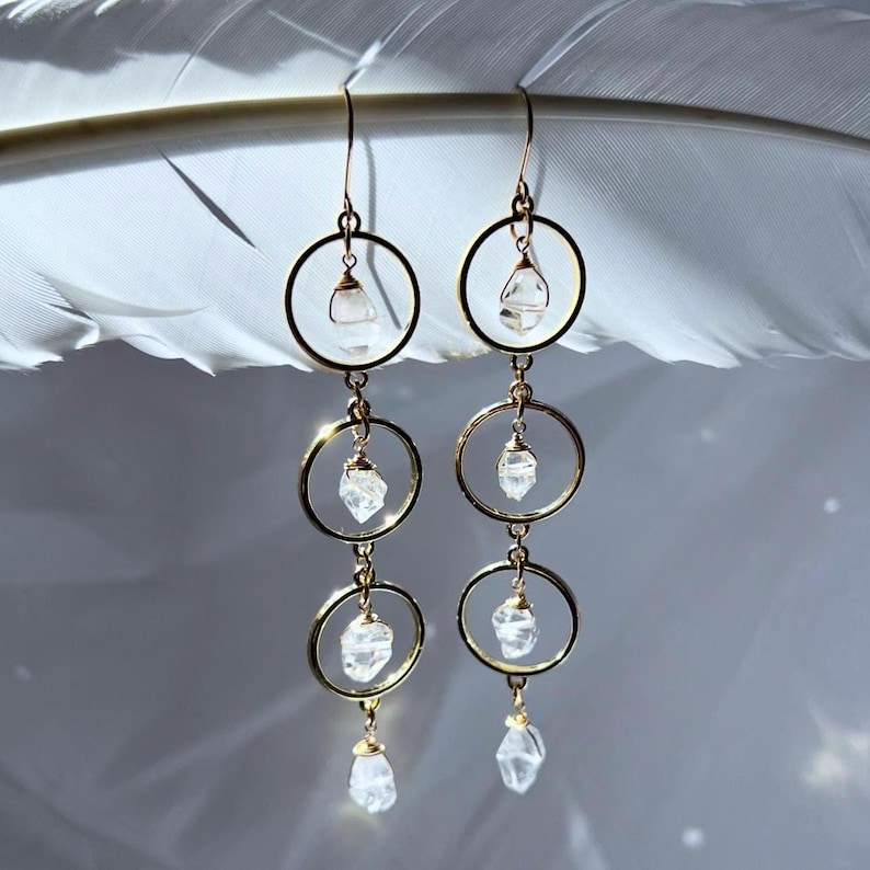 May include: Gold-tone hoop earrings with clear, faceted crystal accents. Each earring features three connected hoops, with a crystal suspended within each hoop and at the bottom. The earrings are displayed against a soft, blurred background.