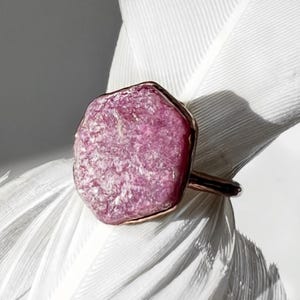 May include: A pink gemstone ring with a rough texture and a copper-toned band. The ring is set against a white background. The irregularly shaped gemstone is the focal point, with its pink hue contrasting the white backdrop.