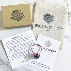 Puede incluir: Un anillo de color cobre con una gema en bruto de color p&uacute;rpura oscuro, presentado con una peque&ntilde;a caja de cart&oacute;n, una bolsa de tela y instrucciones de cuidado. La marca "Buddha Blossom Jewels" es visible en el embalaje.