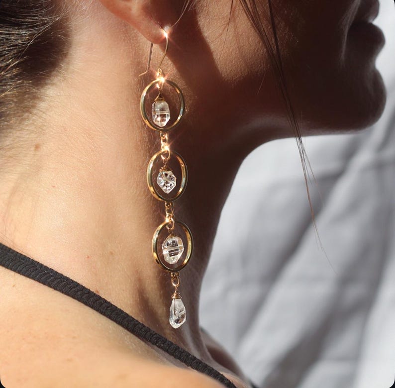 May include: Gold-tone earrings with three connected hoops, each holding a clear, faceted crystal. The earrings dangle from a hook, showcasing a delicate and elegant design. The crystals catch the light, adding sparkle to the jewelry.