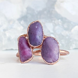 May include: Three copper-colored rings, each featuring a large, irregularly shaped purple gemstone. The gemstones have a natural, raw appearance. The rings are displayed against a soft, blurred background.