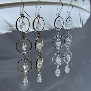 May include: Dangling earrings with clear quartz crystals and gold-colored and silver-colored metal hoops. The earrings have a layered design, with three hoops of decreasing size, each holding a crystal. The earrings are displayed against a white feather.