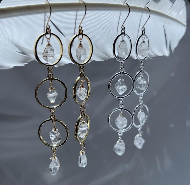 May include: Dangling earrings with clear quartz crystals and gold-colored and silver-colored metal hoops. The earrings have a layered design, with three hoops of decreasing size, each holding a crystal. The earrings are displayed against a white feather.