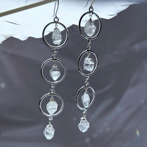 May include: Dangling earrings featuring clear quartz crystals suspended within silver-toned circular frames. The earrings have a layered design with multiple crystal pendants, creating a delicate and elegant accessory. The crystals are faceted, catching the light.
