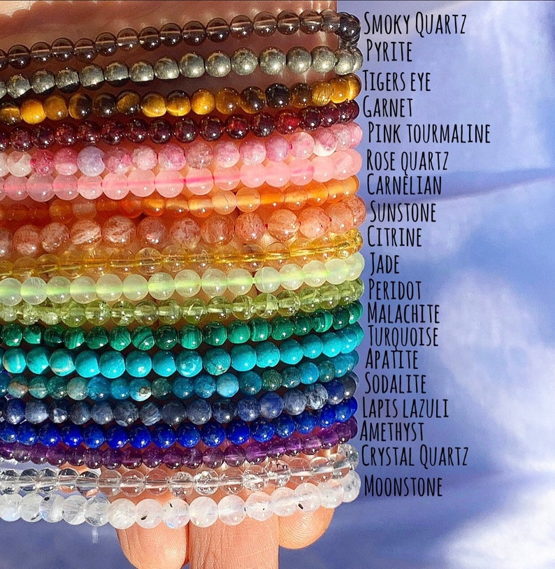 A hand holding 19 strands of different gemstones, in color order to match a rainbow. Beads are 4mm in size and polished, with a round shape. You get topick which crystal calls to you.