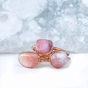 May include: Three rose gold rings with pink opal stones. The rings are stacked on top of each other. The stones are different shapes and sizes.