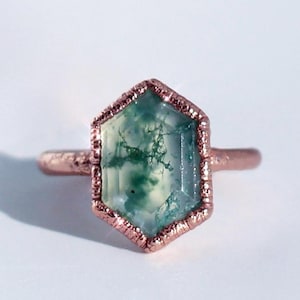 May include: A copper-colored ring featuring a large, hexagonal gemstone. The gemstone is a translucent green with intricate, moss-like inclusions. The ring has a textured bezel setting and a simple band.