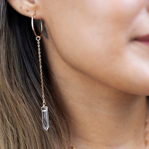 May include: Gold hoop earrings with a delicate chain and a clear quartz crystal pendant.