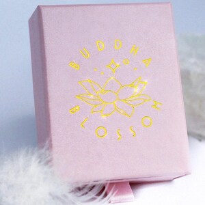 May include: A light pink jewelry box with the words "BUDDHA BLOSSOM" in gold lettering. The box features a gold lotus flower design and a pink ribbon pull. The box is made of a soft, textured material.