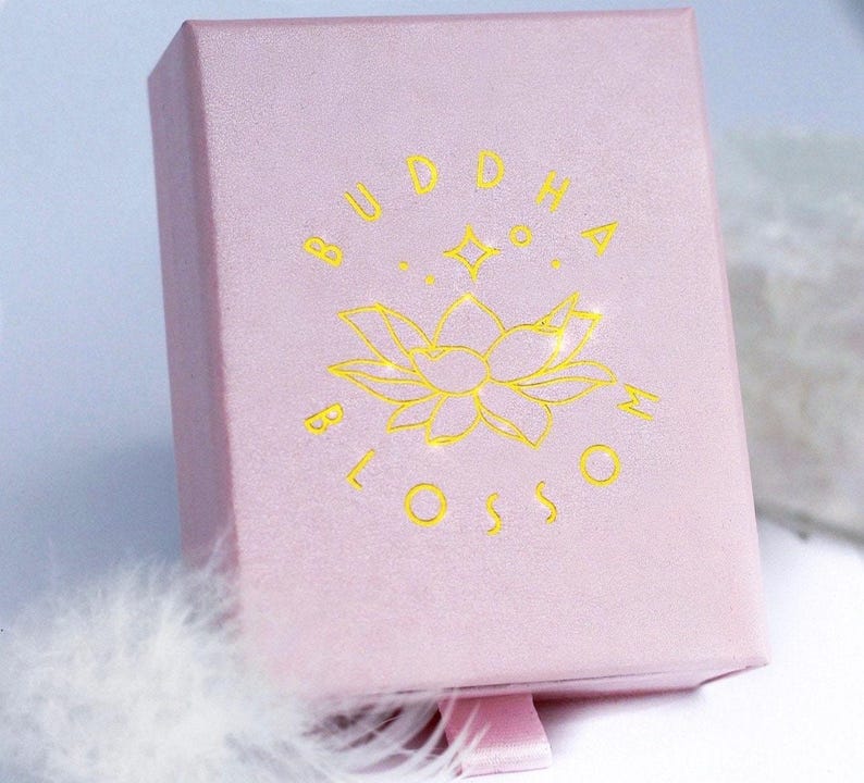 May include: A light pink jewelry box with the words "BUDDHA BLOSSOM" in gold lettering. The box features a gold lotus flower design and a pink ribbon pull. The box is made of a soft, textured material.