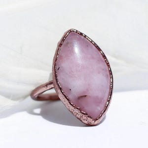 May include: A handcrafted ring featuring a large, oval-shaped, light pink gemstone set in a copper-colored metal band. The gemstone has a smooth surface with subtle variations in color. The metal setting has a textured, rustic appearance.