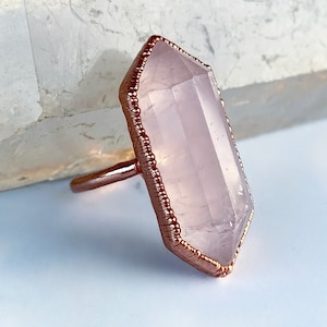 May include: A rose quartz ring with a copper wire wrapped around the stone. The ring is set on a white marble surface.