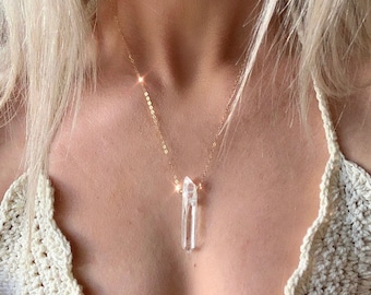 Puede incluir: Un collar de cadena dorada con un colgante de cristal de cuarzo transparente. El colgante es una punta larga y delgada.