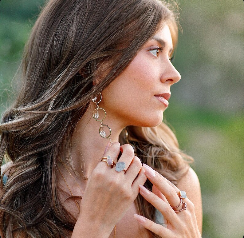 May include: A woman wearing gold-toned jewelry, including long earrings with dangling circles and gemstone accents, and multiple rings. The rings feature various gemstones. The woman has long, wavy brown hair.