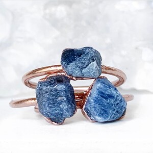 May include: Three raw blue sapphire stones are wrapped in rose gold wire and set on a ring band. The ring is made of copper wire.
