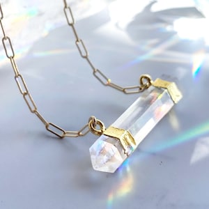 May include: A gold chain necklace with a clear quartz crystal pendant. The crystal is faceted and has a gold trim at each end.