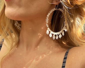 May include: Gold hoop earrings with a dangling crystal design. The crystals are clear and faceted, creating a sparkling effect.