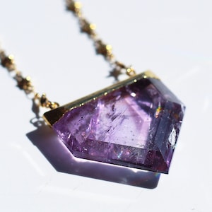 Peut inclure: Un collier en chaîne dorée avec un grand pendentif en améthyste facetté. Le pendentif a la forme d'un diamant et a une teinte violette.