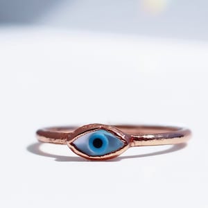 May include: A rose gold ring with a blue evil eye charm. The charm is set in a white shell-like material.