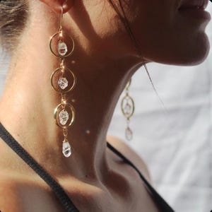 May include: Gold-toned earrings with three circular hoops, each suspending a clear, faceted crystal. The earrings are long and dangle, with a delicate design. The crystals catch the light, creating a sparkling effect. The earrings are a jewelry item.