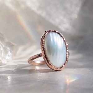 May include: A ring featuring an oval-shaped, iridescent white stone set in a textured, rose-gold colored metal band. The setting has a distinct, rugged edge that frames the smooth, light-reflecting surface of the central stone, creating a contrast.