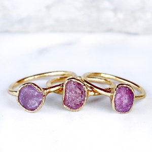 Könnte beinhalten: Drei goldene Ringe mit rosafarbenen Edelsteineinsätzen. Die Ringe sind übereinander gestapelt. Die Edelsteine sind roh und haben ein natürliches, unpoliertes Aussehen.