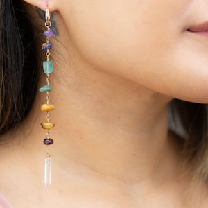 May include: A long, dangling earring with a gold chain and a variety of colorful gemstones, including amethyst, lapis lazuli, green aventurine, tiger's eye, and garnet. The earring ends with a clear quartz crystal.