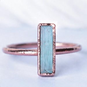 May include: A ring featuring a vertical, rectangular, light blue stone set in a textured rose-colored metal band. The stone is encased in a matching rose-colored metal frame with a slightly hammered or rustic finish along the edges of the band.