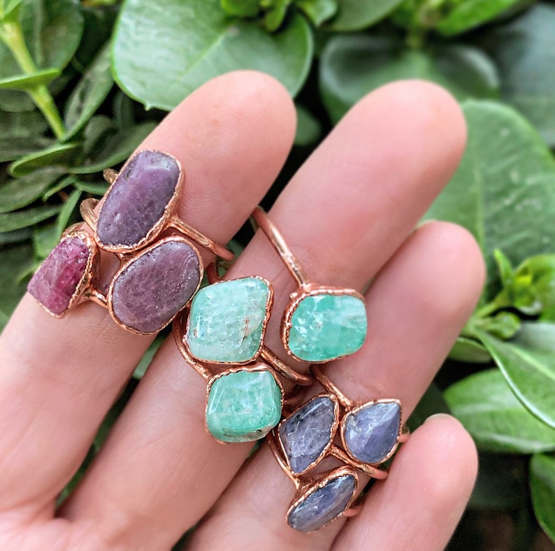 Puede incluir: Una colecci&oacute;n de anillos hechos a mano con piedras preciosas en bruto de varios colores. Los anillos tienen una banda y un engaste de metal color cobre. Las piedras preciosas incluyen tonos de rosa, verde y morado. Fondo de follaje verde.