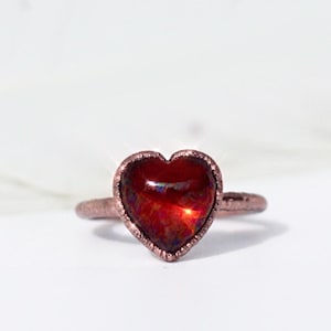 May include: A copper-colored ring featuring a heart-shaped, red gemstone. The gemstone has a textured appearance and is set in a detailed, textured band. The ring is displayed on a white background.