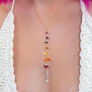 May include: A gold-tone chain necklace featuring a vertical drop design with seven small, multi-colored polished gemstone chips in purple, blue, green, yellow, orange, and red, ending in a clear, faceted, cylindrical crystal point pendant.