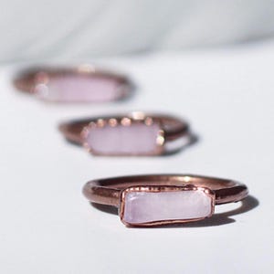 May include: Three rose gold rings with rectangular, light pink stones are displayed on a white surface. The rings are in focus, with the closest ring showing the most detail. The stones have a smooth, polished appearance.