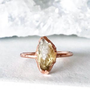 May include: A rose gold ring with a raw, yellow-green crystal set in the center. The crystal has a rough, natural texture and is surrounded by a thin band of metal.