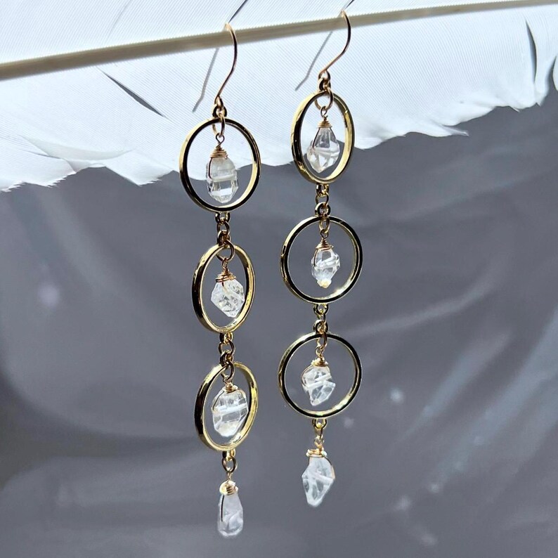 May include: Gold-toned dangle earrings featuring clear quartz crystals and circular frames. The earrings have a delicate design with three quartz crystals suspended within gold hoops, creating a layered effect. The earrings are designed to be worn with a hook.