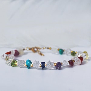 May include: A delicate bracelet featuring a variety of colorful, irregularly shaped gemstones, including clear quartz, turquoise, and amethyst. The stones are strung together with gold-colored beads and a matching clasp. The bracelet is displayed against a soft, white background.