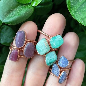 Puede incluir: Anillos artesanales de alambre de cobre con piedras preciosas en bruto de colores. Los anillos muestran piedras moradas, verdes y azules. Los anillos se exhiben sobre un fondo de follaje verde.