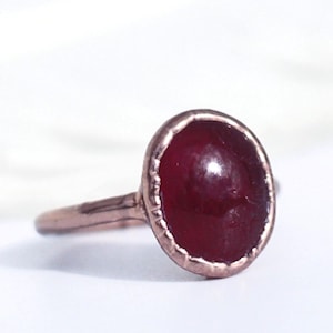 May include: A handcrafted ring featuring a large, oval-shaped, deep red gemstone set in a copper-colored metal band. The ring has a textured bezel setting and a simple, slender band. The gemstone is the focal point, with a polished, glossy surface.