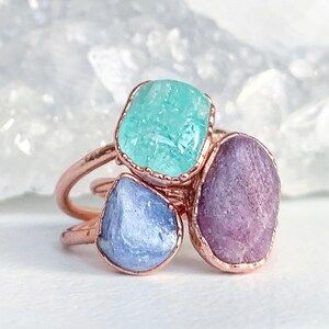 Puede incluir: Anillo de tono cobre con tres piedras preciosas en bruto. Las piedras son de color azul claro, turquesa y morado. El anillo tiene un dise&ntilde;o org&aacute;nico, que resalta la belleza natural de las piedras. Una joya &uacute;nica.