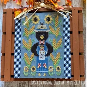 Puede incluir: Patrón de punto de cruz con un cuervo de dibujos animados que lleva un mono y un sombrero, sosteniendo una taza que dice "Fall Y'all". El cuervo está rodeado de girasoles y mazorcas de maíz. El patrón se titula "Clovis and Stanley: A Sip of the Seasons".