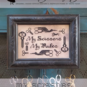 May include: A framed cross-stitch sampler with black thread on white fabric. The sampler reads "My Scissors My Rules" and features a design of scissors and birds. The frame is black with ornate detailing and is hanging on a blue wall with hooks and scissors hanging below.