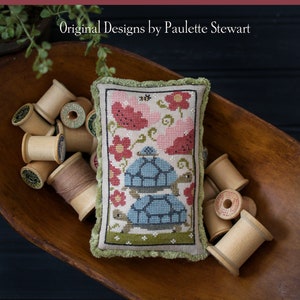 May include: A cross-stitch sampler pillow with a green border and a design of three tortoises stacked on top of each other, surrounded by pink flowers. The pillow is sitting in a wooden bowl with spools of thread. The text "Tortoise Tower" is at the bottom of the image.